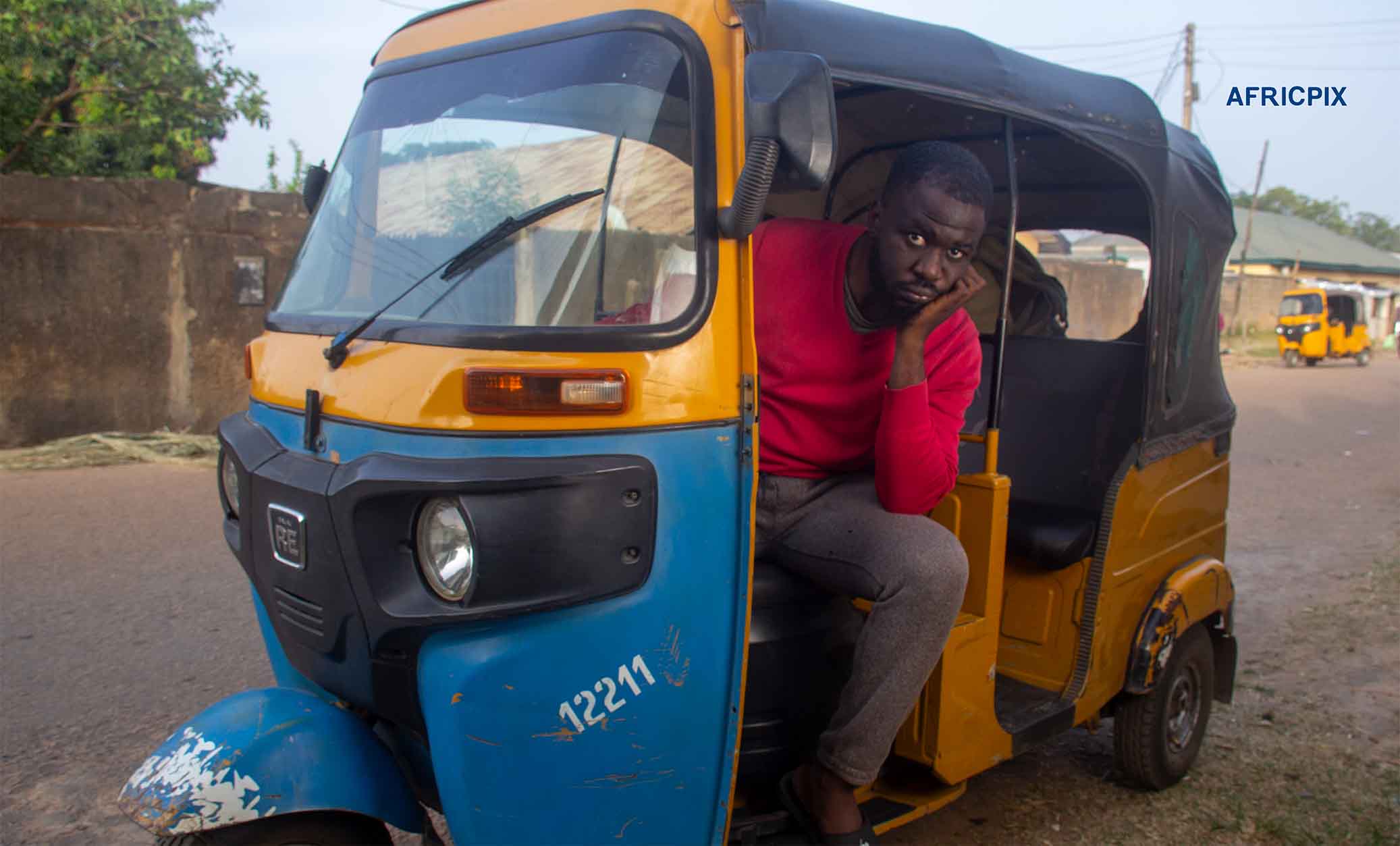 An African tricycle rider sitting inside his tricycle with his hand on his cheek, looking sad and distressed 2.